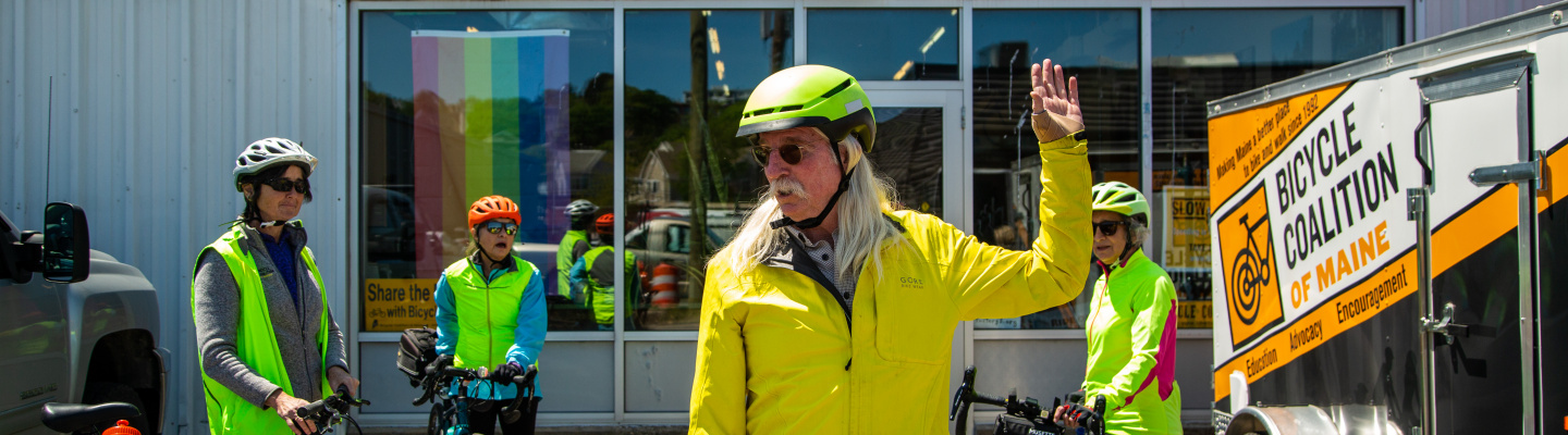 A man in bright yellow teaches bicycling hand signals to a group of cyclists